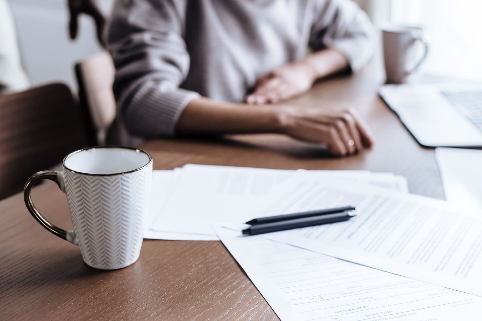 Hands resting on a kitchen table beside paperwork and a cup of tea.